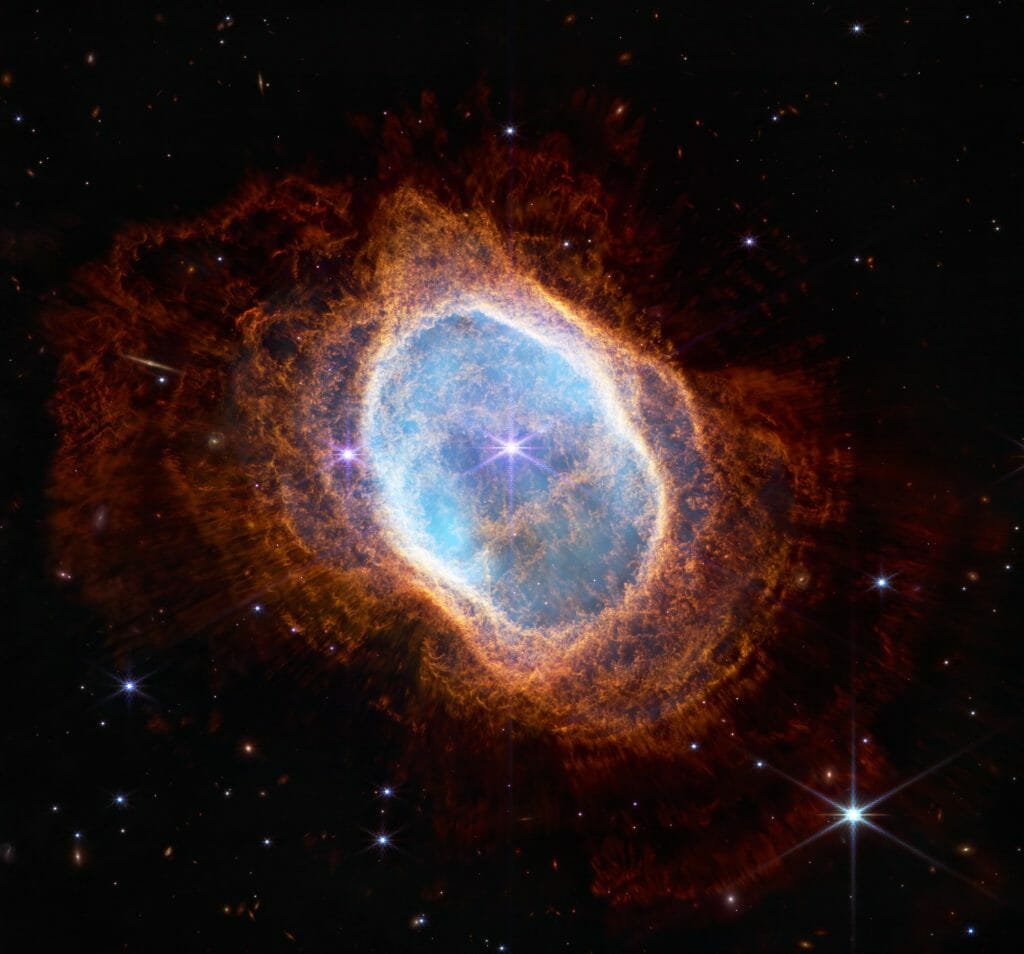 Southern Ring Nebula (NIRCam Image)
Description / Alt-Text
Colorful image of near-infrared light from a glowing cloud with a distorted ring-like shape, illuminated
from within by a bright central star. The Southern Ring Nebula is a large, semi-transparent oval that is
slightly angled from top left to bottom right. A bright white star appears at the center of this image. A
large transparent teal oval surrounds the central star. Several red shells surround the teal oval,
extending almost to the edges of the image. The shells become a deeper red with distance from the
center. The bright central star has eight diffraction spikes. Behind the gaseous teal layers are deeper
orange layers that are arranged like threads in a complex weaving. The red layers, which are wavy
overall, look like they have very thin straight lines piercing through them, which are holes where light
from a central star is traveling. The background of the image is black and speckled with tiny bright stars
and distant galaxies.