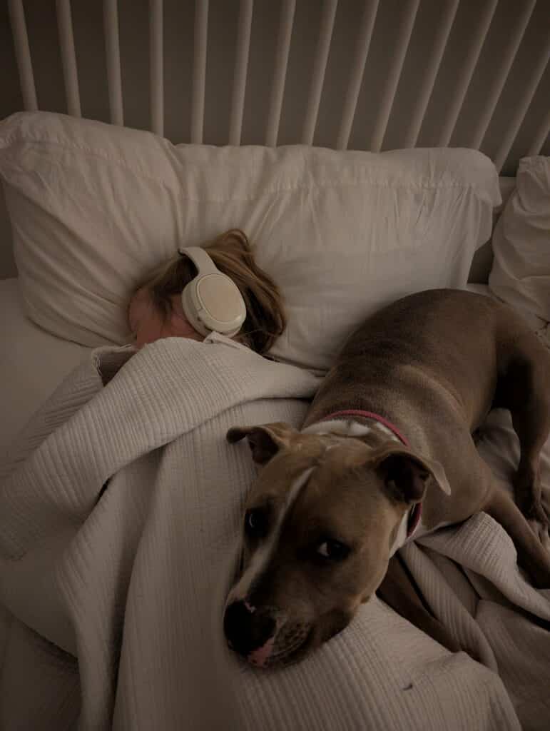 A little girls sleeps with headphones on under the covers while Stola, a little brown pit bull lays on top of her to snuggle. 