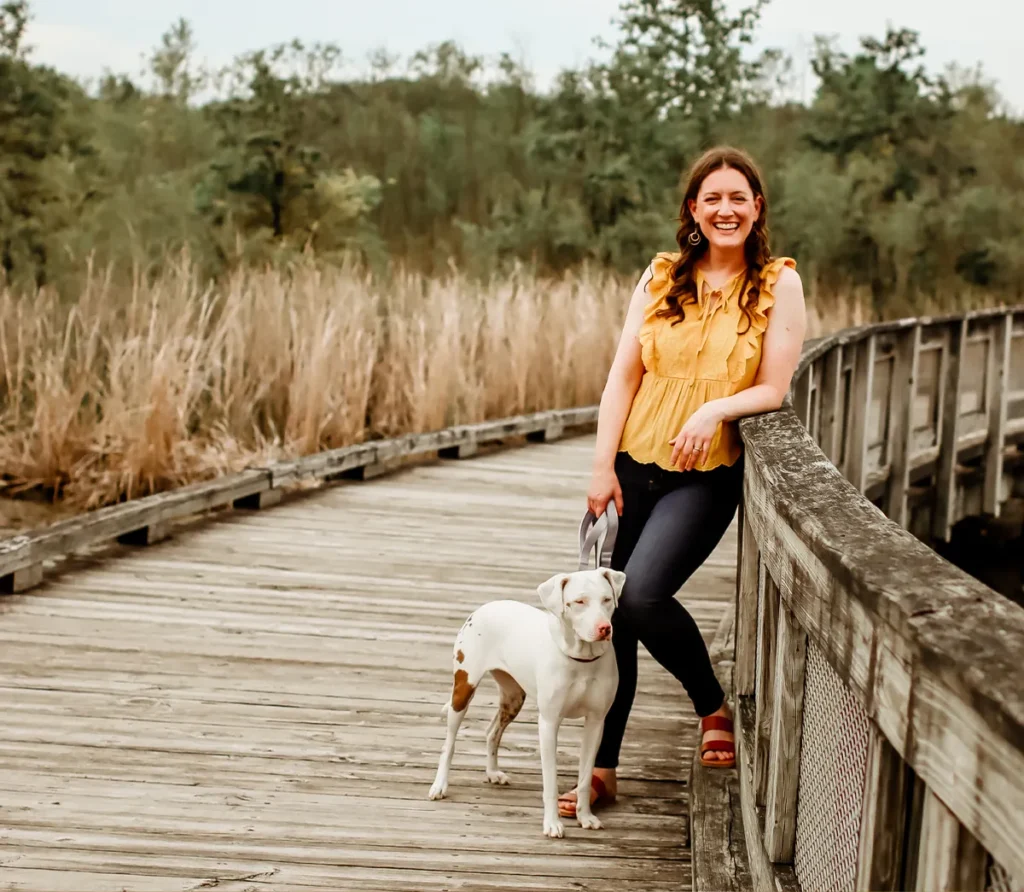 The author wears a yellow blouse and black denim jeans with brown sandals and stands on a weathered bridge holding the purple leash of a mid-sized white dog with brown speckles on her hind end. 